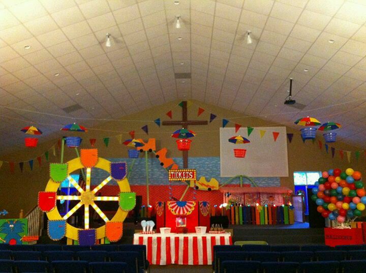 an indoor carnival themed party with colorful decorations and balloons on the ceiling, in front of a large screen