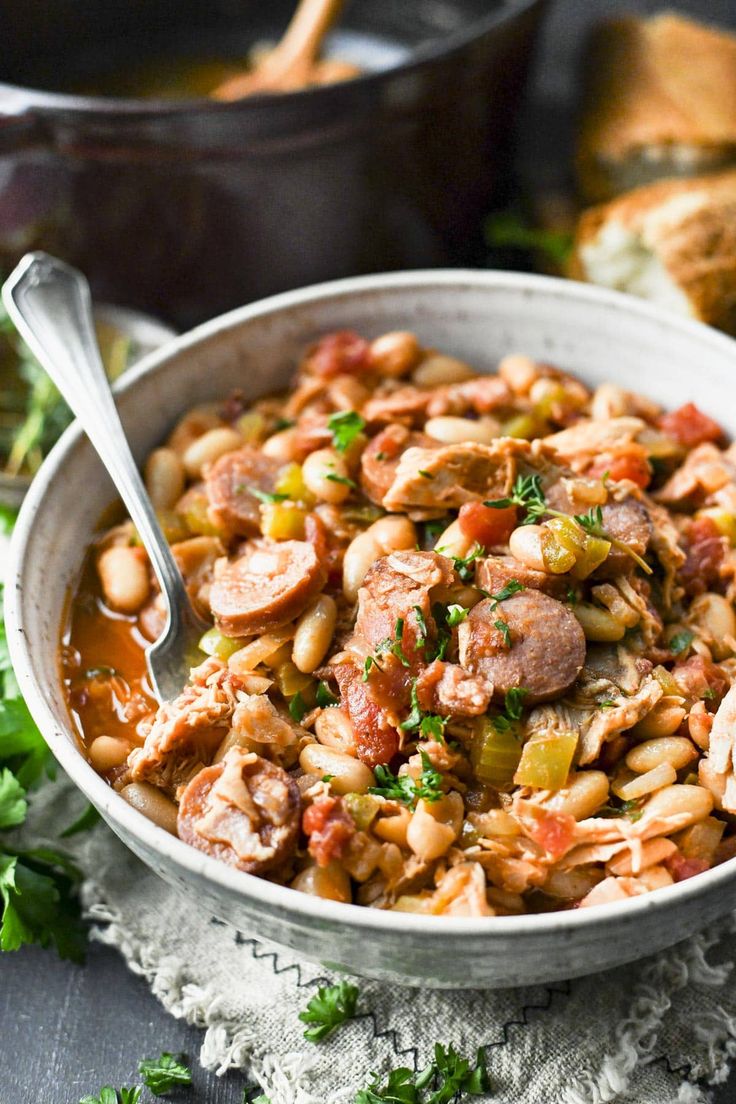 a bowl filled with beans and sausage next to some bread on a tablecloth,