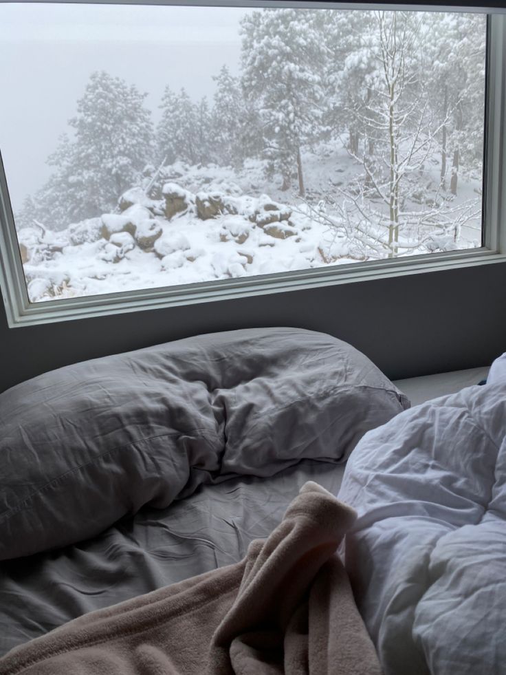 an unmade bed in front of a window with snow on the ground and trees outside