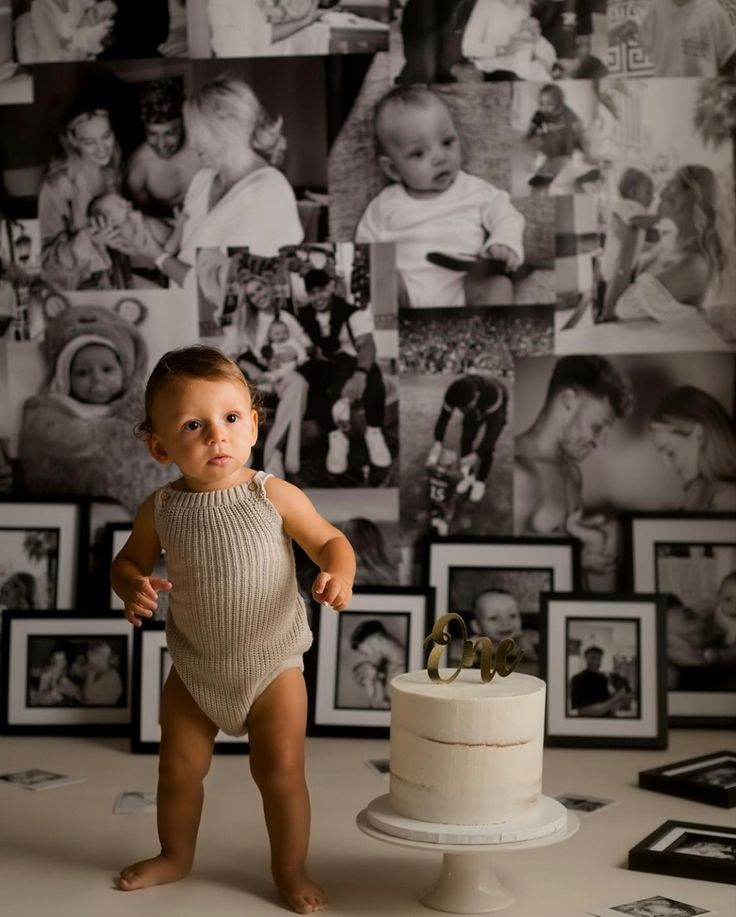 a baby standing in front of a cake with pictures on the wall behind it,