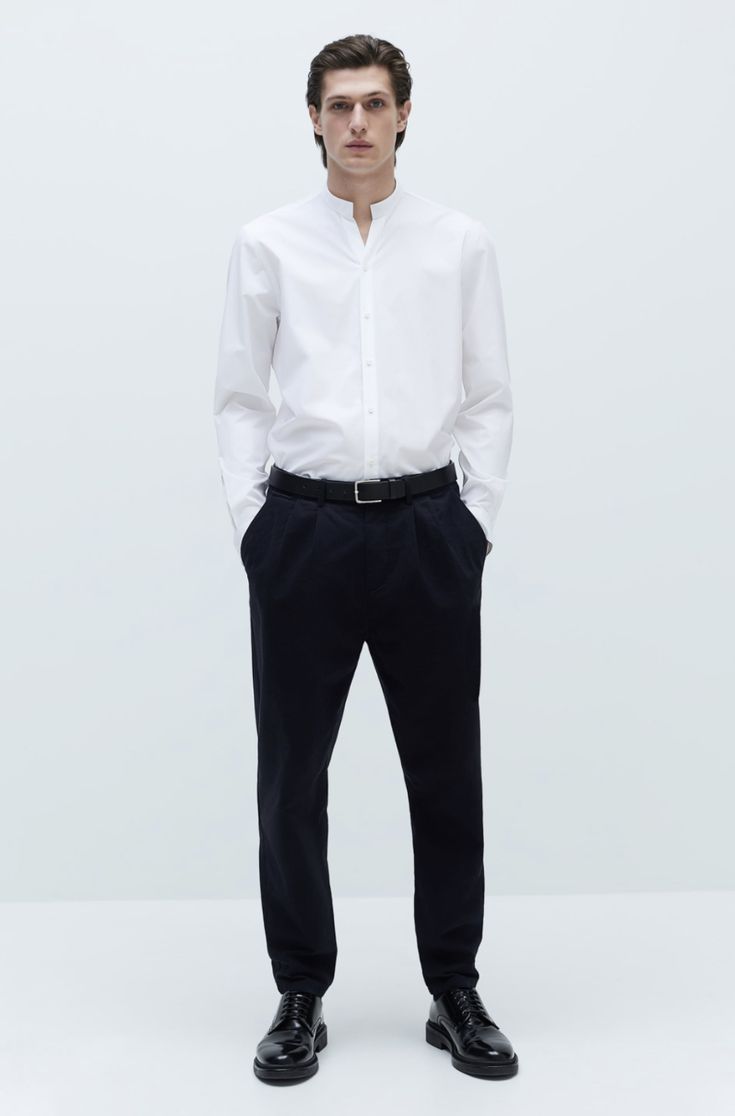 a man standing in front of a white wall wearing black pants and a white shirt