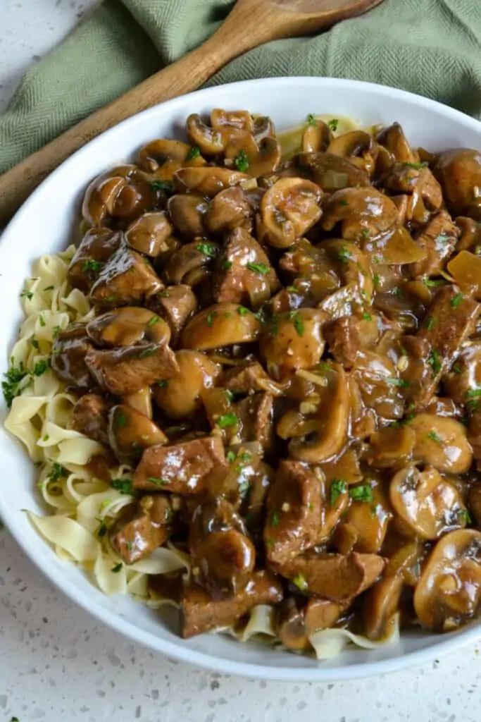 a white bowl filled with pasta covered in meat and mushroom sauce next to a wooden spoon