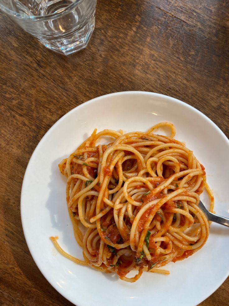 a white plate topped with spaghetti on top of a wooden table next to a glass of water