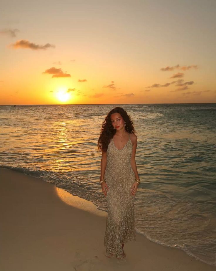 a beautiful woman standing on top of a sandy beach next to the ocean at sunset