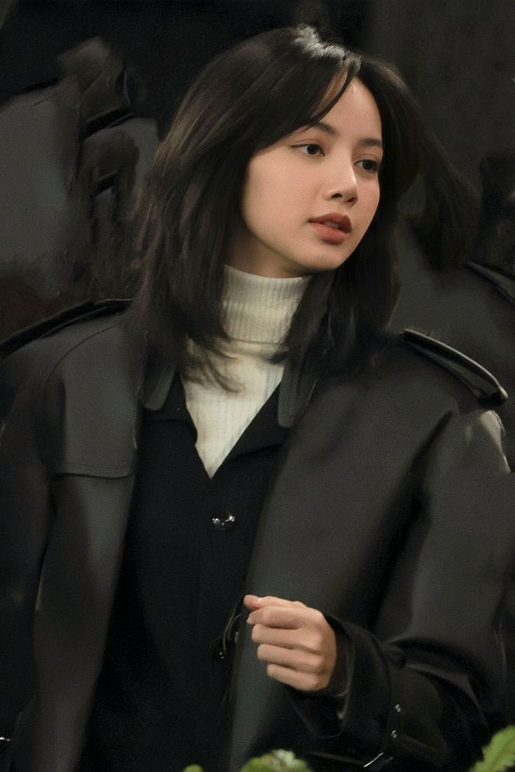 a woman with long black hair wearing a coat and turtle neck sweater standing in front of plants