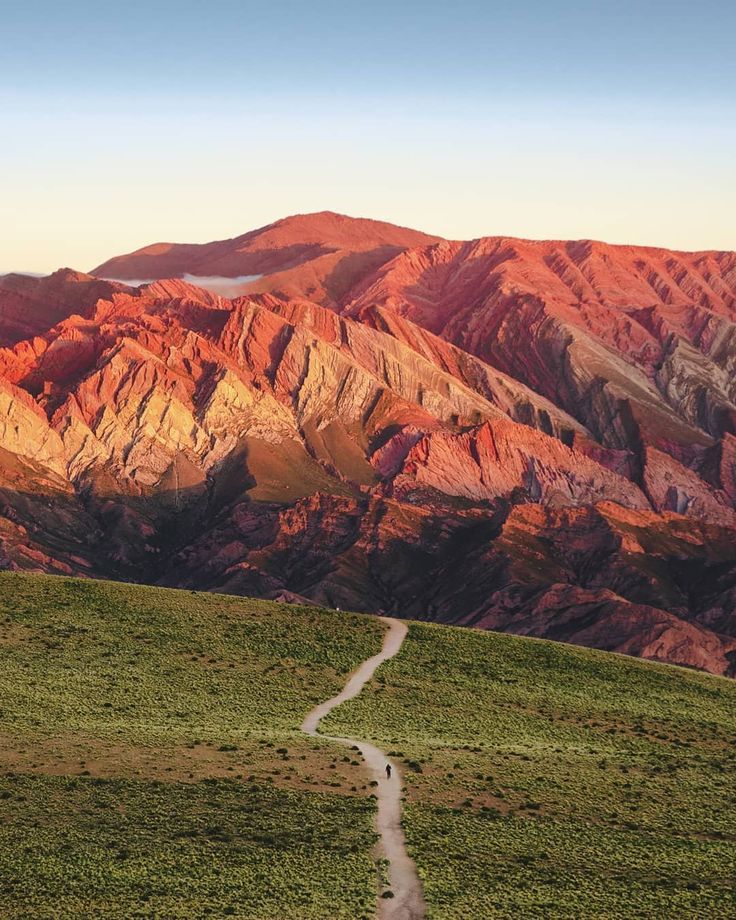 a dirt road in the middle of a grassy field with mountains in the background at sunset