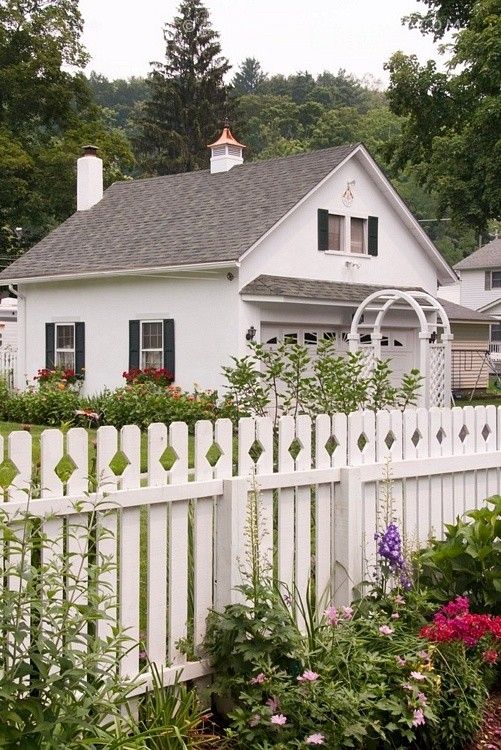 a white picket fence in front of a house with lots of flowers and trees around it