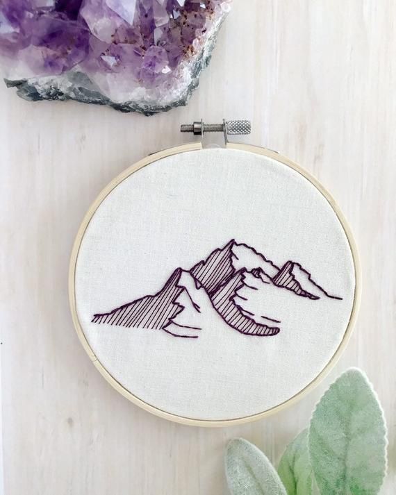 a hand embroidered mountain scene is shown on a wooden table next to some plants and rocks