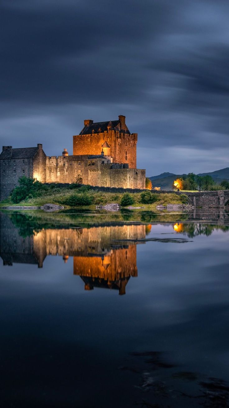 an old castle is lit up at night with its reflection in the water and dark clouds