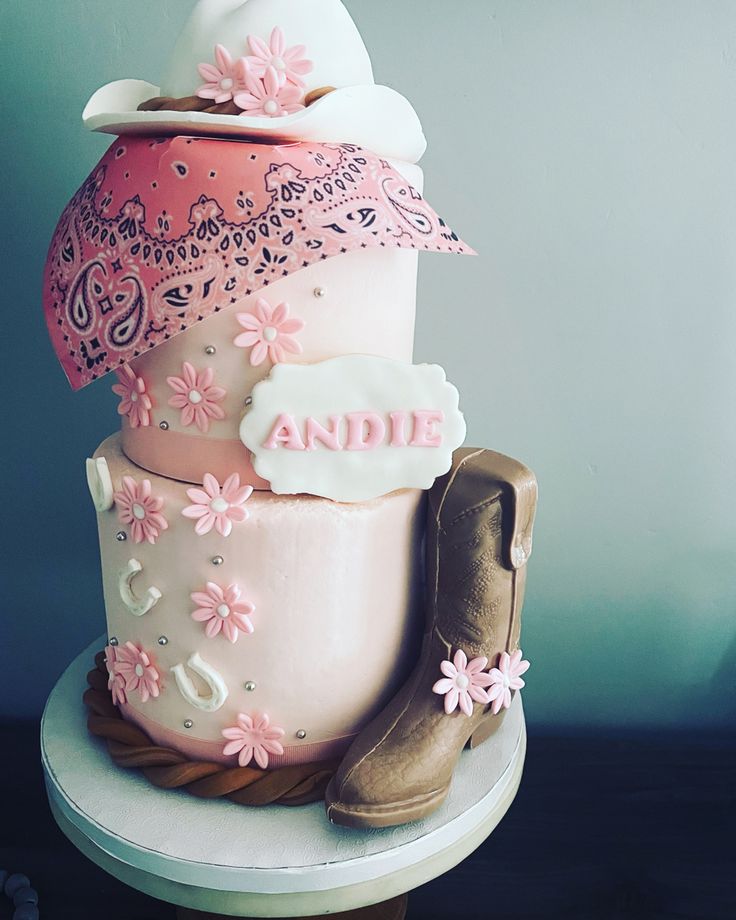 a three tiered cake decorated with pink and white flowers, cowboy boots, and an umbrella