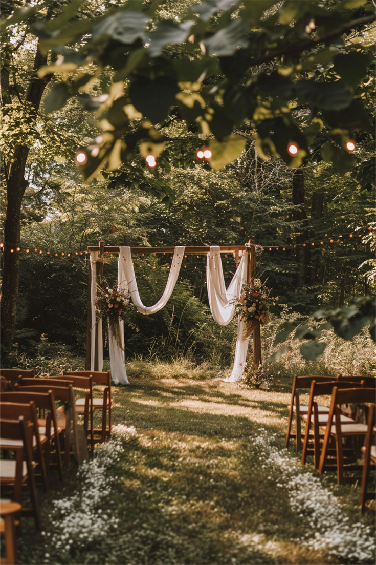 A beautiful garden wedding setup with a wooden arch draped in white fabric and adorned with floral arrangements, surrounded by string lights and wooden chairs under tall trees. Ceremony In The Woods, Hay Bell Wedding Seats Chairs, Fall Wedding Trellis Ideas, Nature Wedding Arch, Bohemian Vintage Wedding, Backyard Wedding Small Space, Wedding Alters Outside, Small Wedding In The Mountains, Wedding Ideas Field