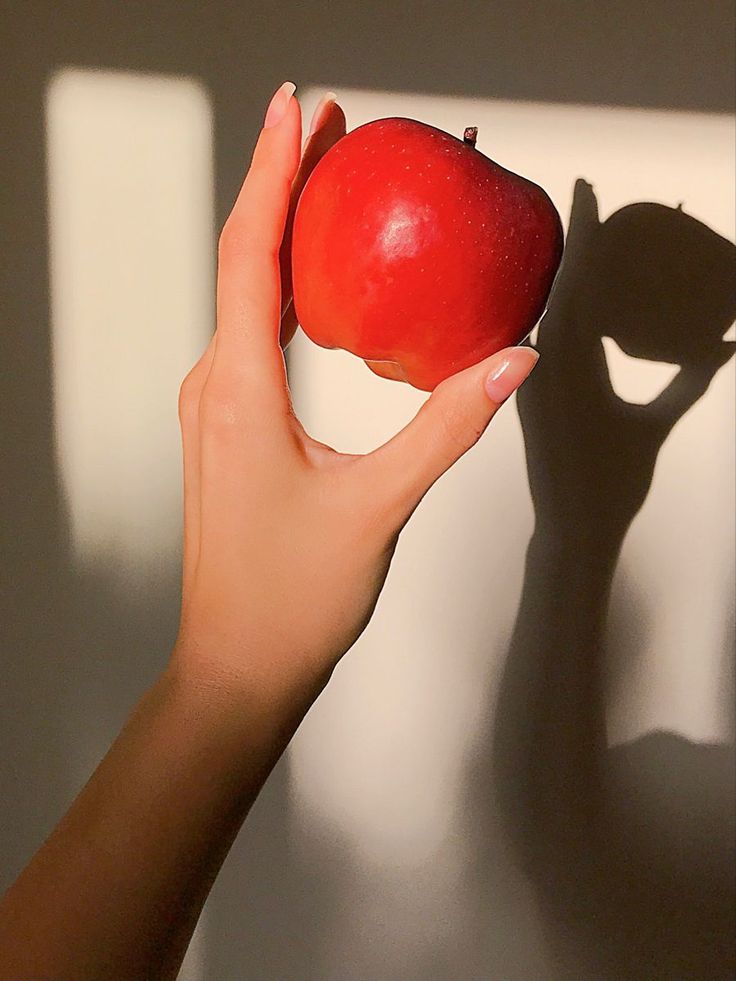 a woman's hand holding an apple in front of the shadow of a wall