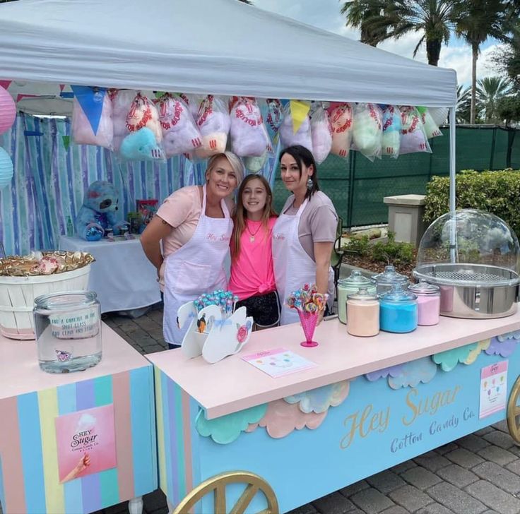 three women are standing behind a pink and blue ice cream vendor's table with decorations on it