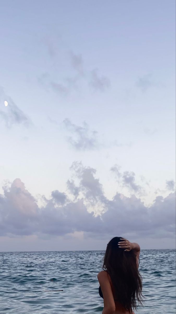 a woman standing in the ocean with her back to the camera and looking at the sky