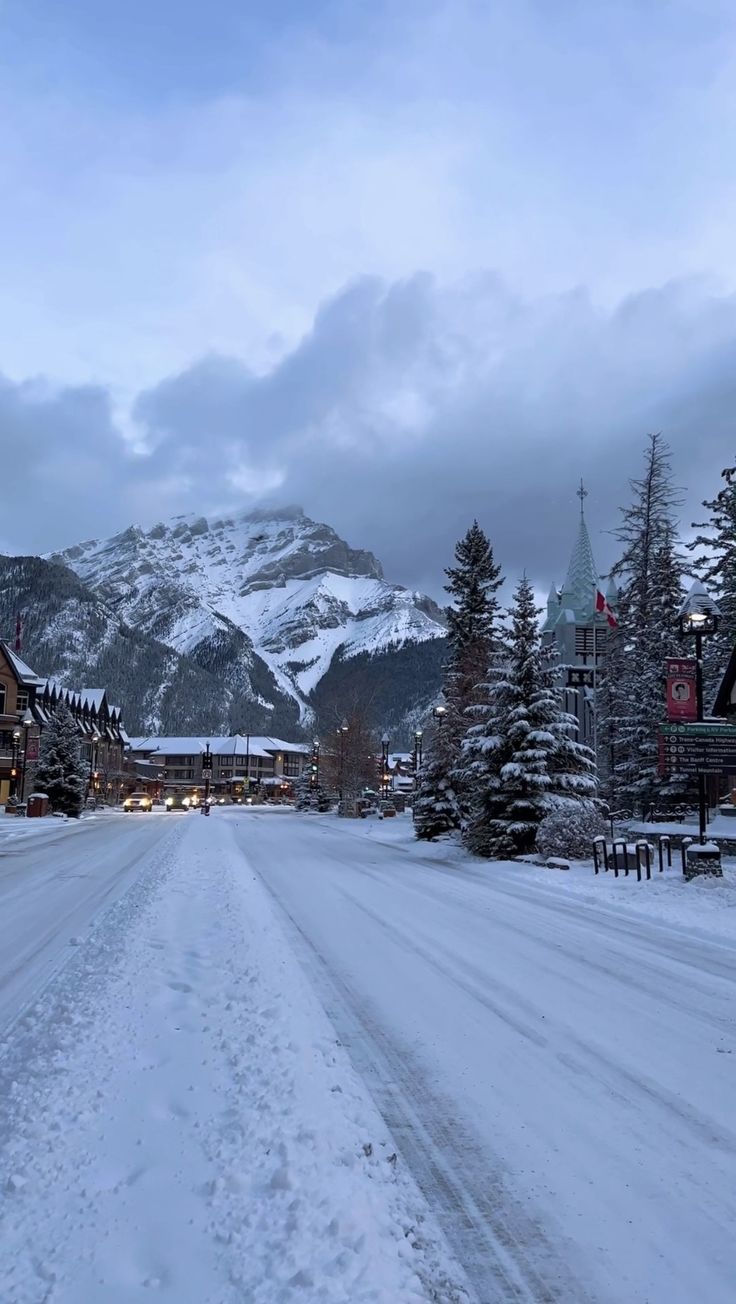 a snow covered street with buildings and mountains in the background on a cloudy day at dusk