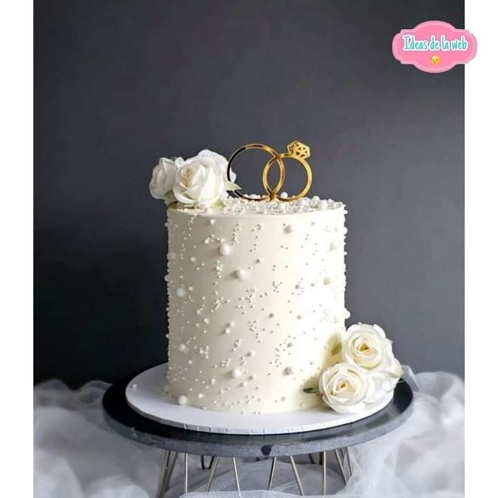 a wedding cake with white flowers and two gold rings on top, sitting on a table