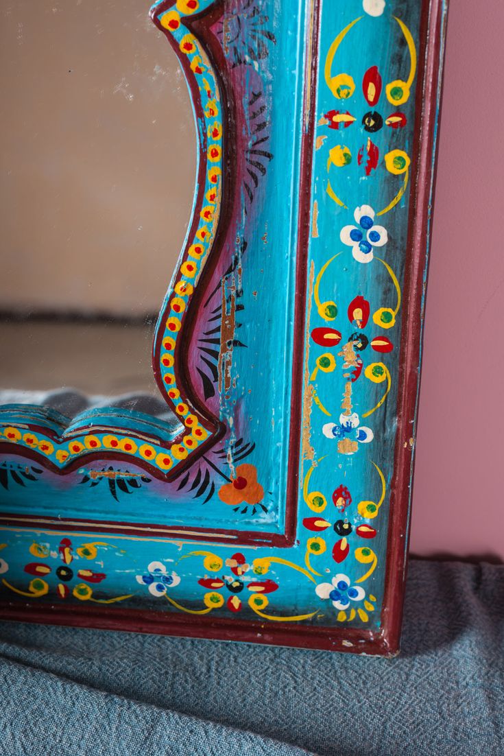 an ornate blue and red mirror with flowers on it's sides, in front of a pink wall