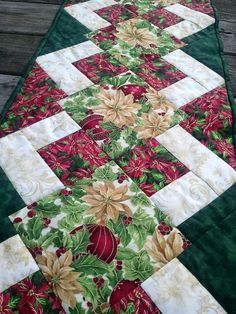 a green table runner with red, white and green flowers on the top is sitting on a wooden deck