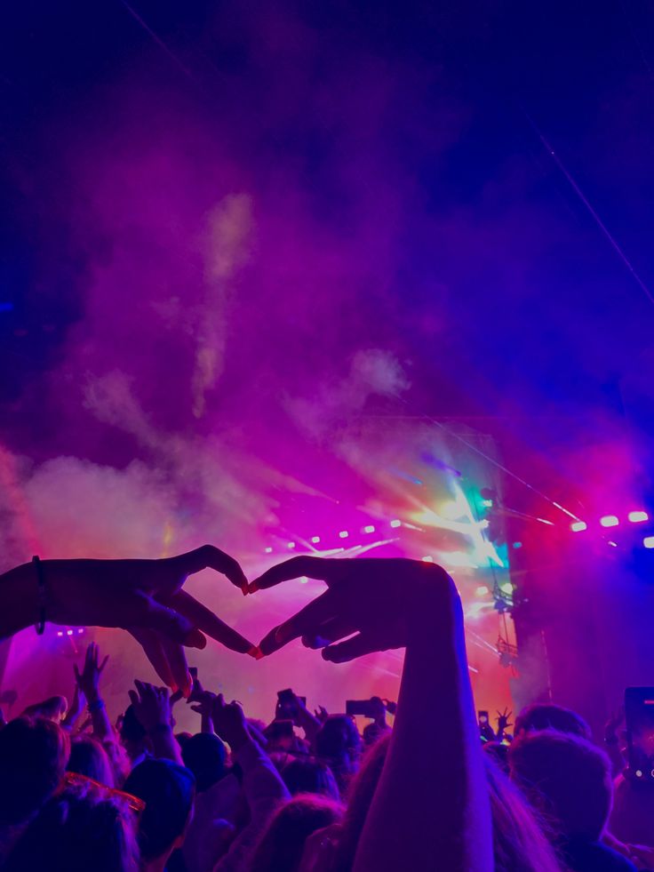 two hands making a heart shape in front of a crowd at a music festival with colored lights