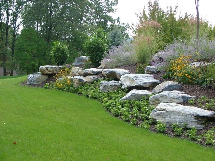 a large rock wall in the middle of a lush green field with flowers and trees