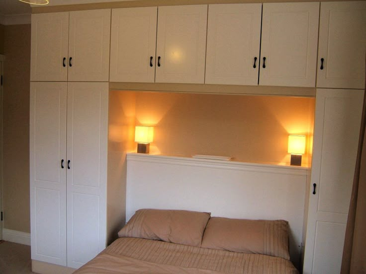 a bed sitting under two lamps in a bedroom next to white cupboards and drawers