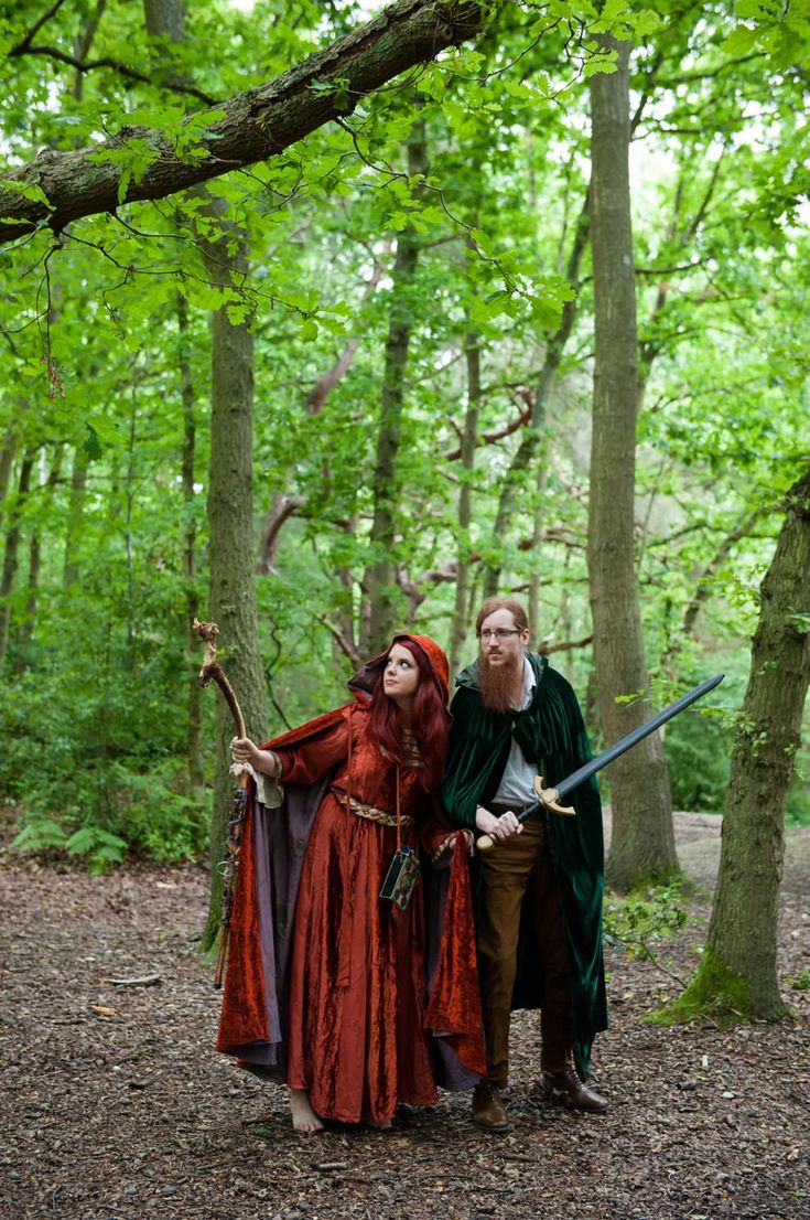 two people dressed up in costumes standing next to each other on the forest floor with trees behind them