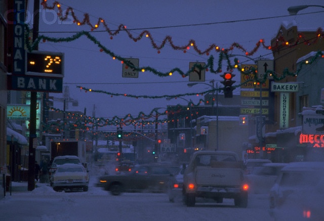 cars driving down a snowy street with christmas lights strung over the buildings and traffic signs