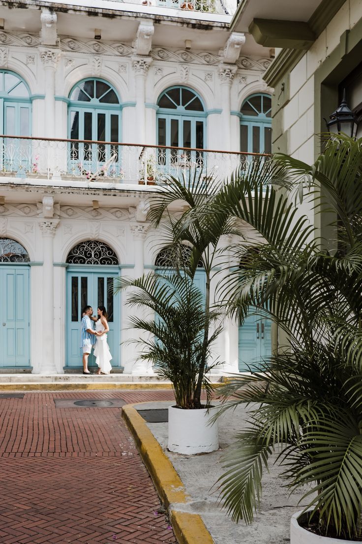 two people standing in front of a white building with blue shutters and palm trees