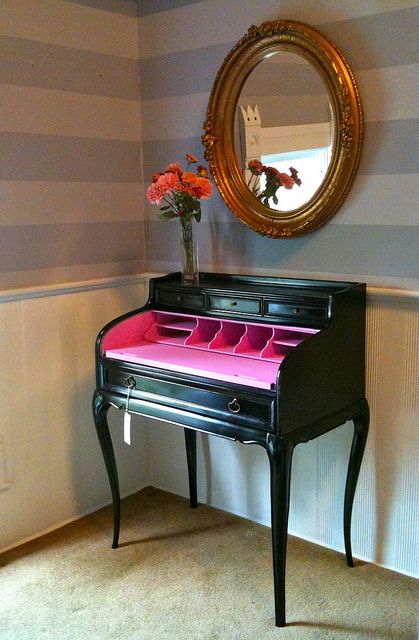 a small black desk with pink drawers in front of a mirror and striped wallpaper