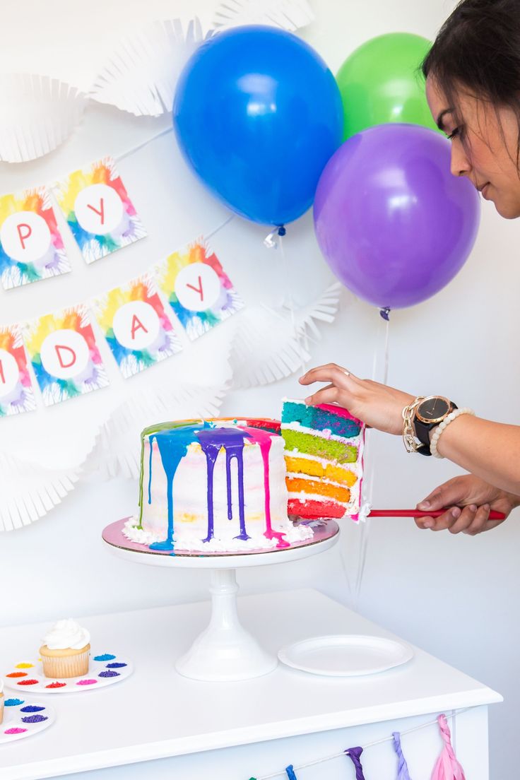 a woman is decorating a birthday cake with colorful icing and balloons in the background