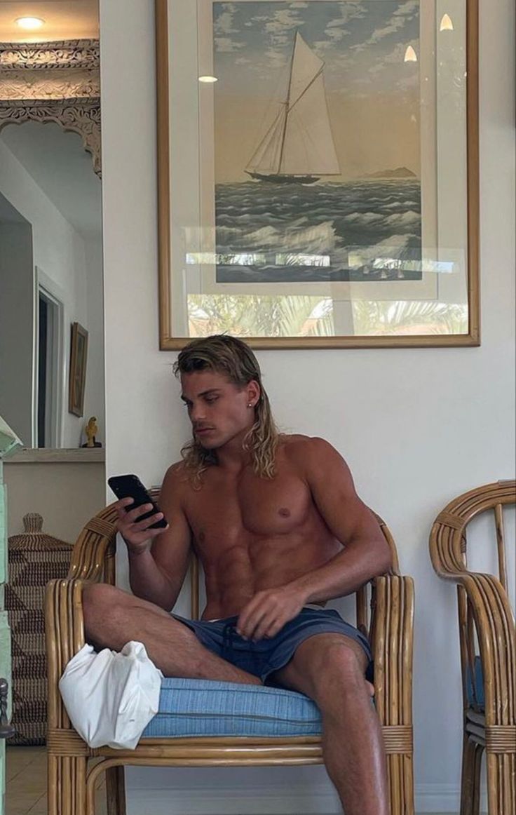 a shirtless man sitting on a chair looking at his cell phone in the living room