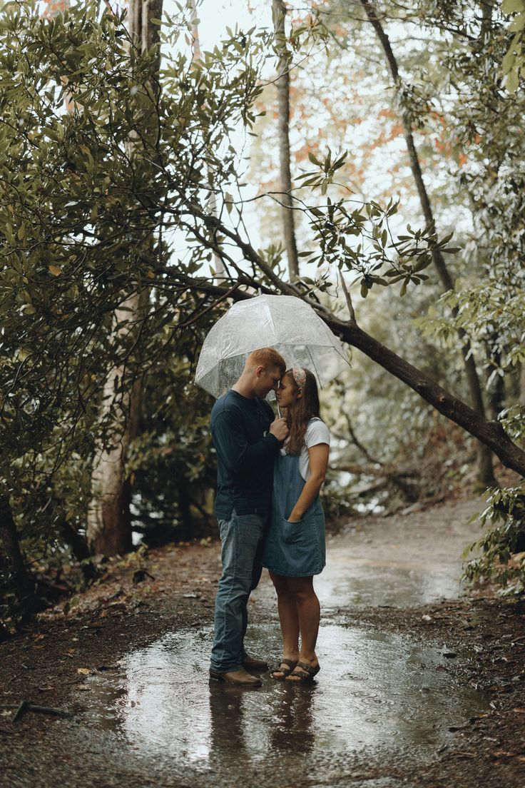 a man and woman standing under an umbrella in the rain on a path surrounded by trees