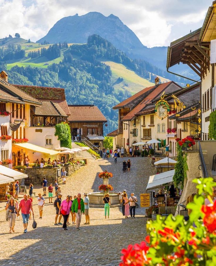 many people are walking down the cobblestone street in an old european town with mountains in the background