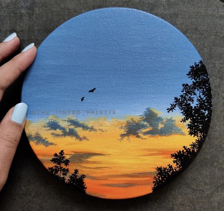 a hand holding up a painted disc with trees and clouds in the sky at sunset