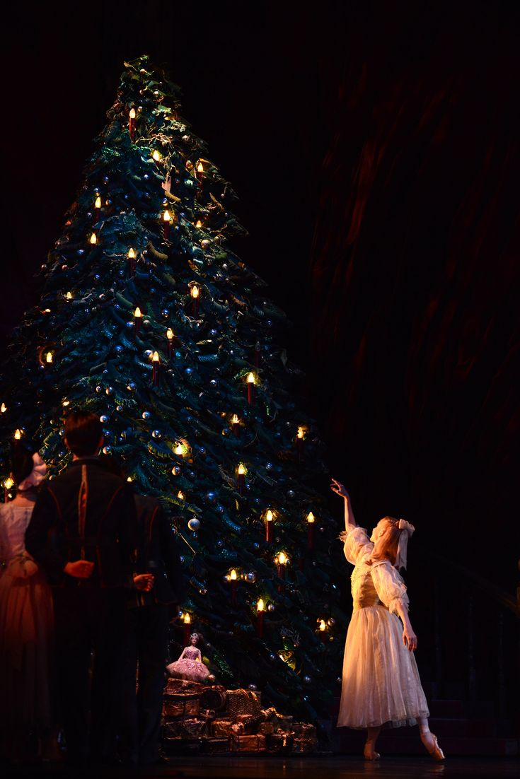 a woman in white dress standing next to a christmas tree with lights on it at night