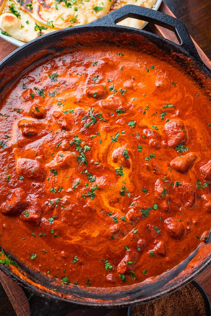 a skillet filled with meat and sauce on top of a wooden table next to bread