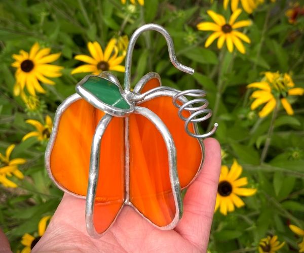 a hand holding an orange stained glass pumpkin ornament in front of yellow flowers