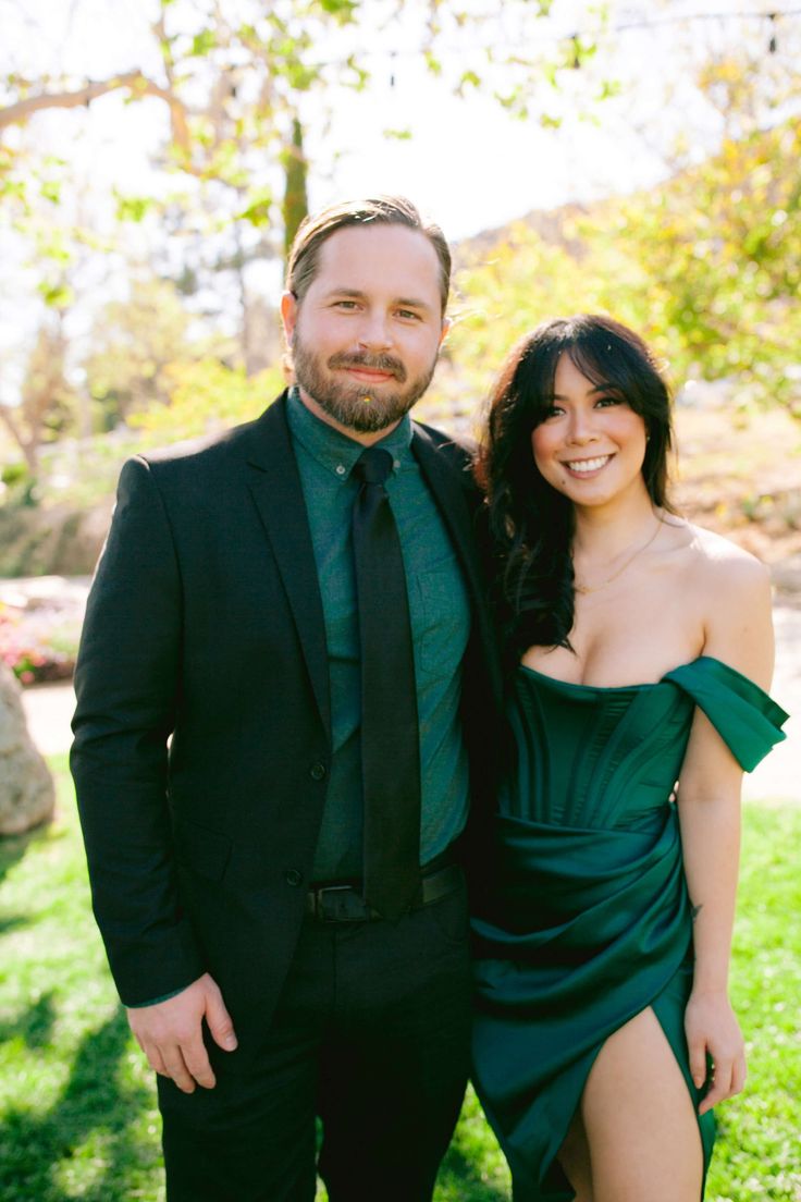 a man and woman in formal wear posing for a photo together on the grass with trees in the background