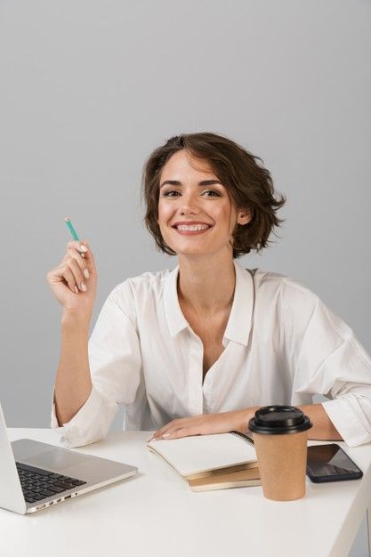 a woman sitting at a desk with a laptop and notebook in front of her, holding a pen