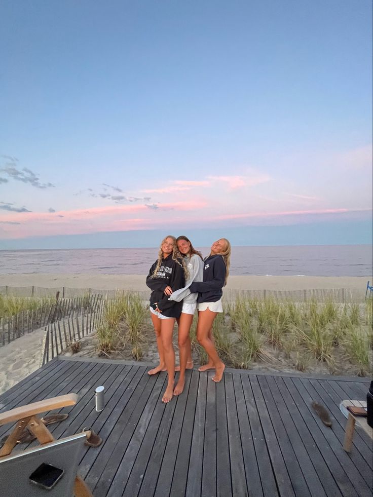 three girls are standing on a boardwalk near the beach at sunset or dawn with their arms around each other