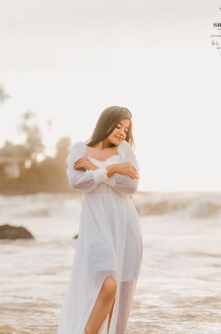 a woman standing in the ocean with her arms around her chest, wearing a white dress