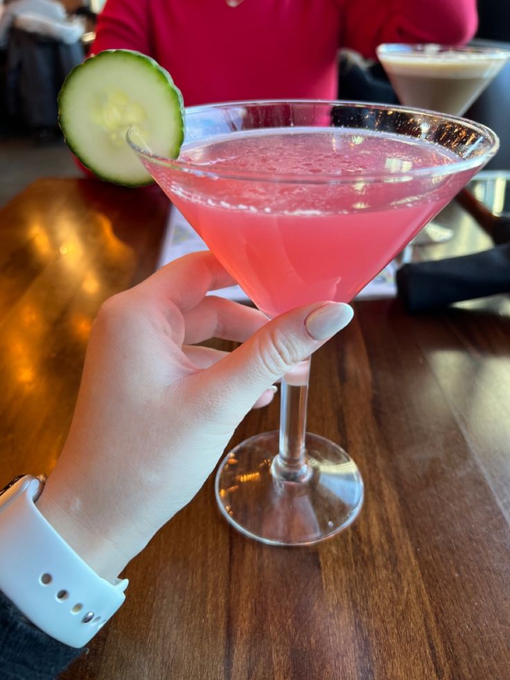 a person holding up a pink cocktail with cucumber on the rim in front of them