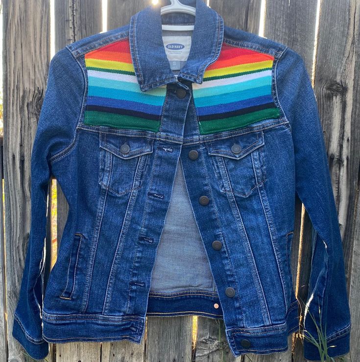 a denim jacket with rainbow stripes on the sleeves and back, hanging against a wooden fence