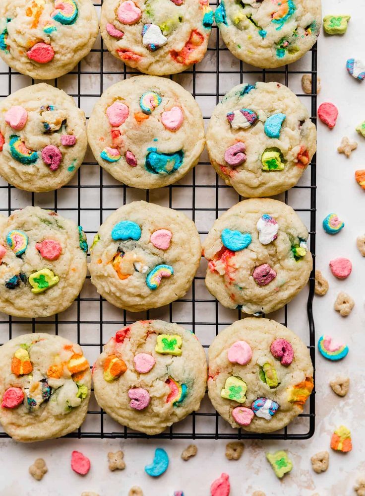 cookies with sprinkles and colored candies are on a cooling rack, surrounded by scattered cereal