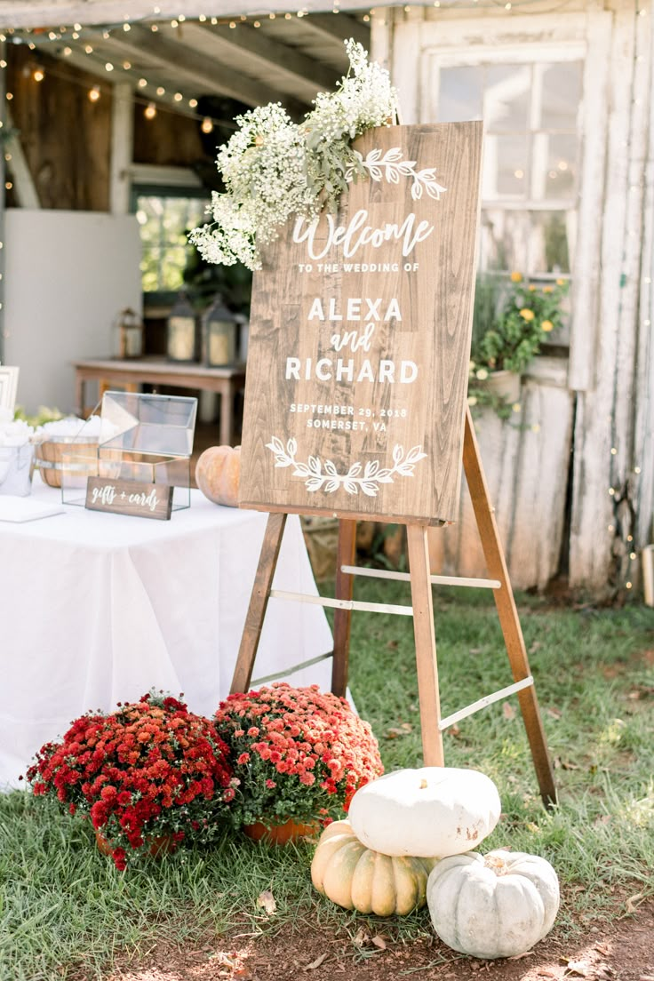 a wooden sign sitting on top of a grass covered field next to flowers and pumpkins