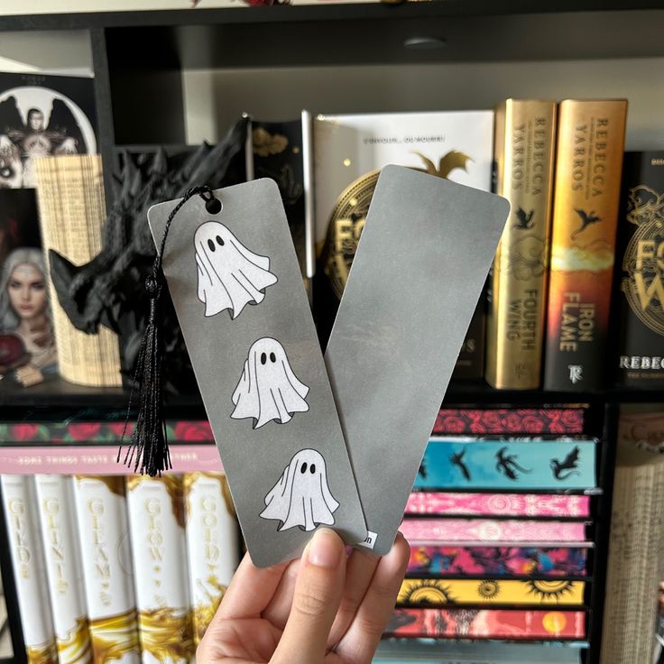 a person holding up two bookmarks with ghost stickers on them in front of bookshelves