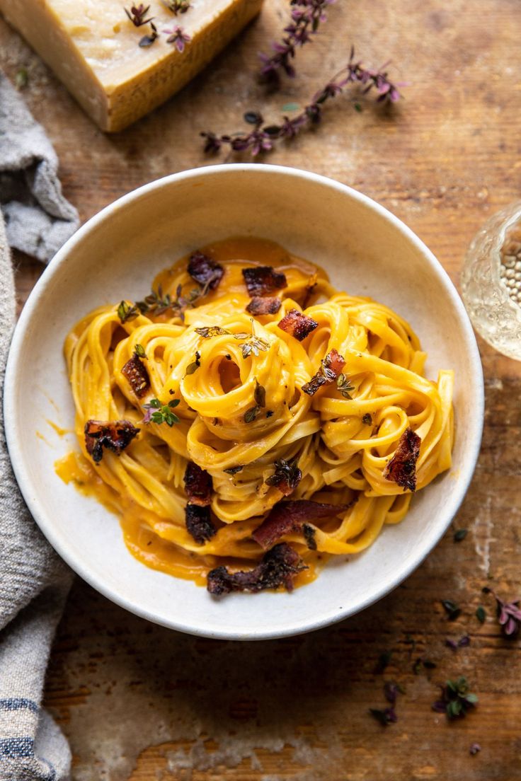 a white bowl filled with pasta and bacon on top of a wooden table next to bread