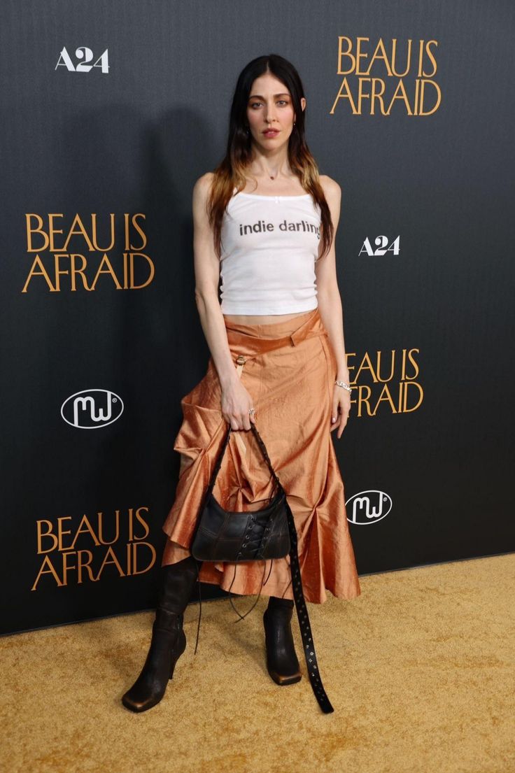 a woman standing in front of a black wall wearing a white tank top and tan skirt