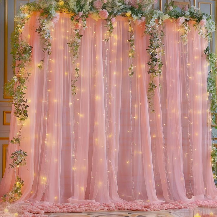 a pink backdrop with flowers and greenery on the top is lit up by fairy lights