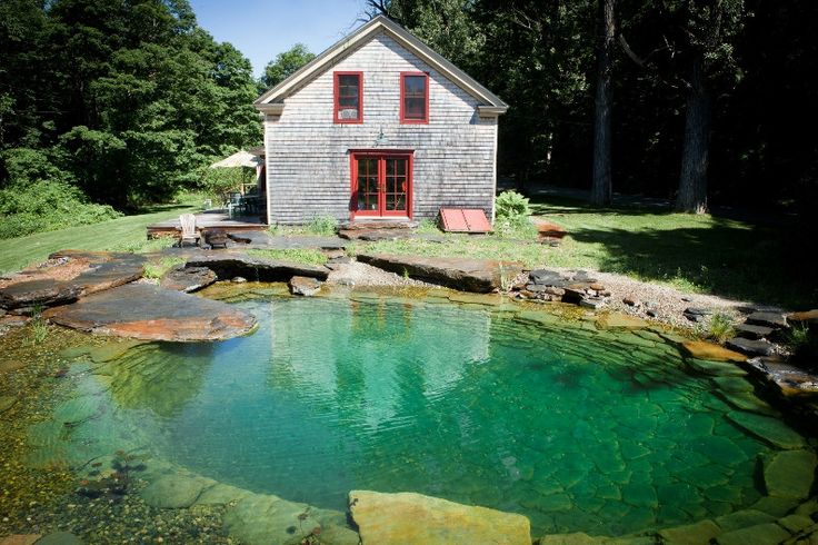 a small house sitting on top of a lush green hillside next to a lake in front of a forest